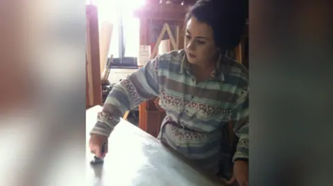 Roisin Quinn Roisin Quinn, who has dark hair in a ponytail, is painting a wooden board in a workshop to create the fake bar. 