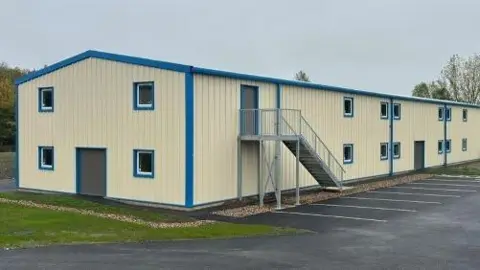 RMBC/Green Belt Experts A cream, warehouse-style building with blue-painted window and door frames. An external stairway leads up to the first floor, with car parking immediately below the stairs and a patch of lawn to the left.