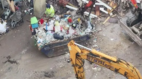 Environment Agency One large skip with rubbish overflowing out of it. The skip is on concrete ground and there is a lot of other rubbish all around it 