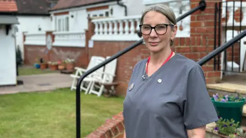 Maureen Lewis, wearing a grey tunic and red lanyard, standing in the garden of St Ives Lodge care home. She has her hair tied back and is wearing glasses.