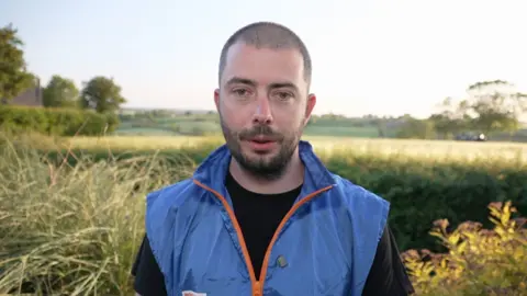 Yevgen has dark cropped hair and a beard and wears a black t-shirt with a blue gilet over it. He is standing in a field.