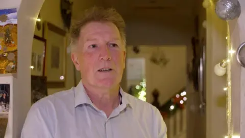 An older man, standing in a doorway in his home. He has light ginger hair and is  wearing a pale shirt with the top button undone. A staircase and Christmas lights can be seen in the background. 