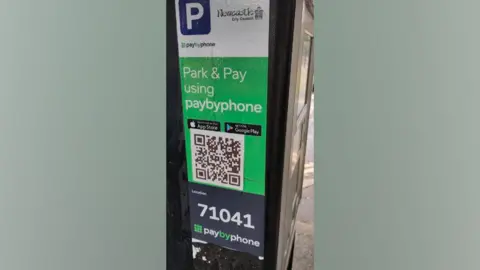 Durham County Council Sticker placed on a parking meter displaying a QR code. The sticker uses the official logos of Newcastle City Council and parking ticket company PaybyPhone. It also includes logos for the Apple App Store and Google Play.