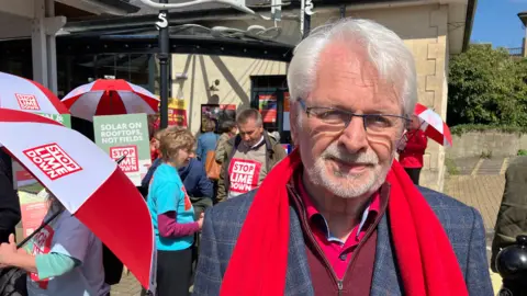 A man smiling softly while wearing a red scarf. Behind him people stood in t-shirts which say 'Stop Lime Down'