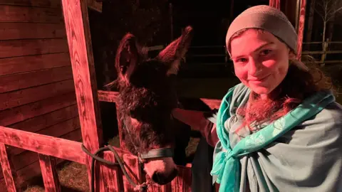 John Devine/BBC Lucy Leeder, 18, has long brown hair, with a skull cap on her head, she has a green shawl on with a green scarf on top she is standing next to a stable, with a donkey.