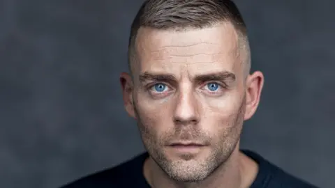 A publicity photo of actor Jamie Dorrington. He has short light brown hair, light blue eyes and stubble on his face.