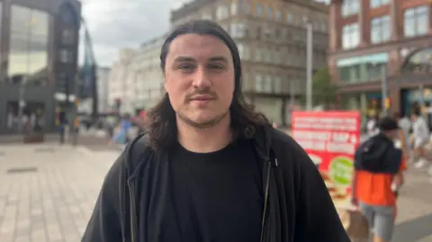 A man with long dark hair, a black t shirt and a black hoodie. He pictured standing in a city street - the background is blurred. 