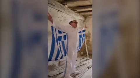 Alan Carr wears a white jumpsuit with the Greek flag attached to both arms