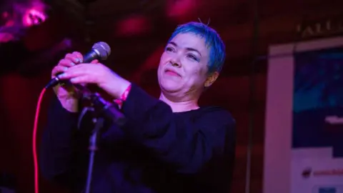 Getty Images Leslie Rankine has short hair which is either dyed blue or lit up blue by stage lights. The rest of the image is lit in red. She is about to sing into a microphone