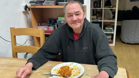 King's Arm Project A man in a room, wearing a black top with a red top underneath. He has a plate of food in front of him that is half eaten, He is sitting at a table, with a chair next to him and items behind him. He is smiling and looking straight at the camera. 