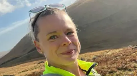 Claire Browne is taking a selfie in front of what looks like a mountain or a hill. She is wearing a fluorescent jacket and has sunglasses perched on her head. She is smiling for the camera.