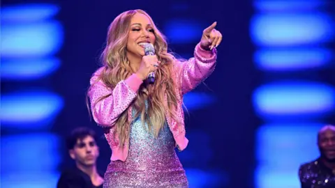 A woman in a pink jacket. She is performing on stage. There are male performers in the background. 