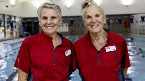Two 80-year-old women stand in front of a swimming pool. Alison, on the left, has short grey hair and is wearing a red short-sleeved sports top. She has a white, rectangular name badge on the left of her top. Jenny, on the right, has longer grey hair tied in a ponytail. She also wears a red short-sleeved sports top, and has a white, rectangular name badge in the same position as Alison's. Both women are smiling.