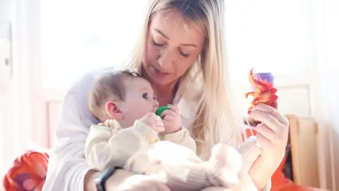 Hayley O'Sullivan holding one of her young daughters. Mrs O'Sullivan has long blonde hair and is wearing a white shirt. She is holding a red toy dinosaur in her left hand. Her baby is wearing a cream outfit and has a green dummy in her mouth.