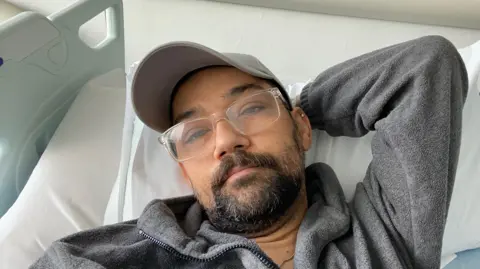 Stephen Hart A man lying in a hospital bed with one arm behind the head, wearing glasses, a grey cap and a grey fleece jacket.