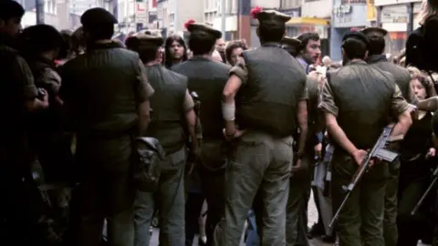 Getty Images It is 1972. Soldiers stand with their backs to the viewer. One of the soldiers on the right of frame is carrying a large calibre gun. Faces of people in the crowd beyond the soldiers can be seen. In the background, a city centre street.