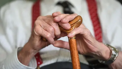 A close up of the hands of an elderley gentleman who is holding onto a walking stick. 