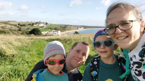 Emilia Shaw A woman with the two boys as well as a man with short hair. A windswept coastline is in the background and the boys are wearing beanie hats and sunglasses.