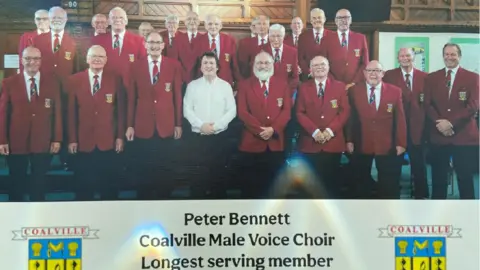 Supplied Choir members stand grouped together in red jackets. They are all smiling. 