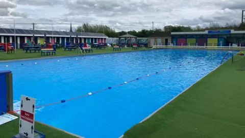 A photo showing an outdoor swimming pool with colourful lane markers. The pool is surrounded by green matting and there are a number of benches and beach huts in the background