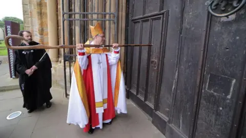 The new Bishop of Durham in his white, red and gold clerical wear holding aloft a long wooden crook which he is using to knock on a large wooden door. Behind him is a church official in long black robes.