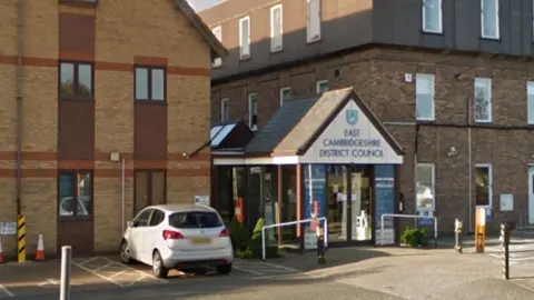 Google 2 buildings are in the picture. One is on the left, and one is on the right. In the middle is a small building which is an entrance to the council building. It has a sign above which says 'East Cambridgeshire District Council'. 