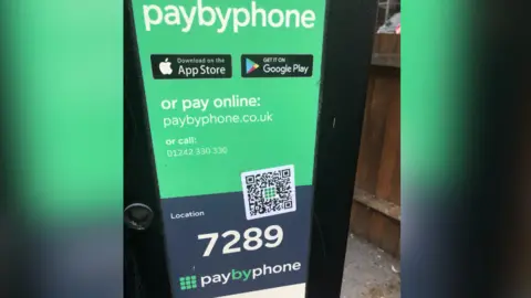 Cheltenham Borough Council A car parking payment machine with a prominent green paybyphone sign at the centre. A location code is displayed in the middle, but a fake QR code sticker is also placed over part of the sign.
