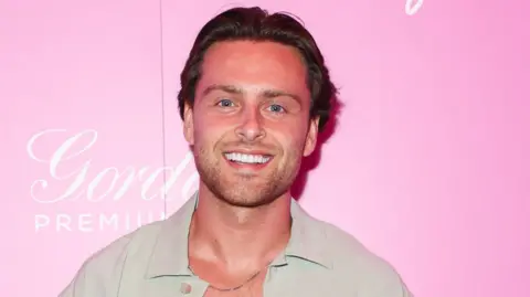 Casey O'Gorman is smiling and stood against a pink background