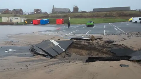 Shane Stanbridge The sinkhole in a tarmac car park. The hole is about as large as three parking spaces. The area surrounding the hole has broken sections of tarmac and sand around it. One side of the car park is covered in water. A handful of cars are parked some distance away from the hole.