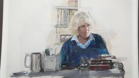 A portrait of Queen Camilla. She is sitting at a desk with a pile of books in front of her. There is a jar of pens and a tankard. She is wearing a blue stripy shirt and blue sweater. There is a book shelf in the background.