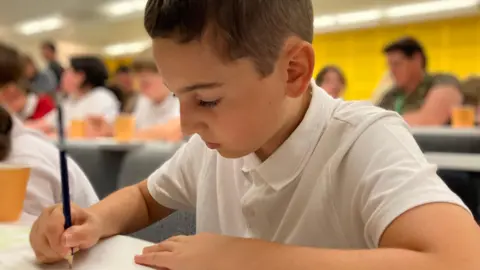 A 10-year-old boy in a white T-shirt is engrossed in writing with a pencil and paper. You can see other children in the background.