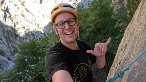 Simon Mutter A man with glasses wearing a black shirt and an orange helmet climbing a rock. He is smiling at the camera and holding up a "hang loose" hand sign.