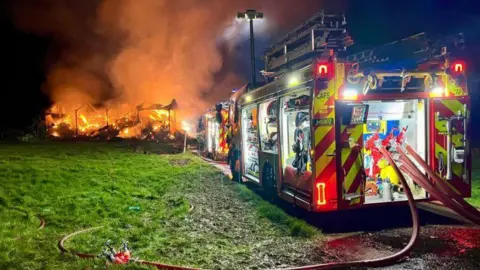 Two fire engines stand next to the blazing barn at night. Fire hoses are laid out on grass nearby.