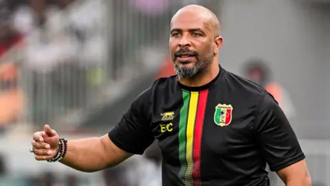 Eric Chelle wears a predominantly black Mali training top, with green, yellow and red stripes on the front and his initials on it as he stands on the touchline during a football match