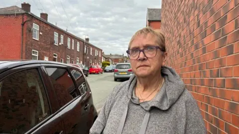 Anne Wells-Bourne has short blonde hair, dark purple spectacles and wears a grey hoodie. She stands next to a parked car on one side and a brick wall on the other. In the background cars are parked on a street of terraced houses.
