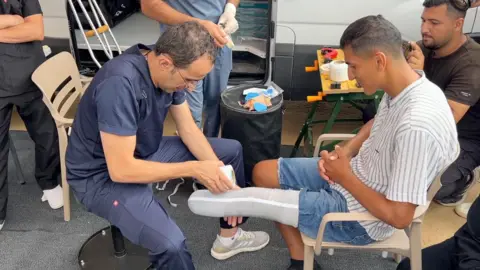 Rizeq Tafish sits on a chair while a Jordanian doctor wraps the stump of his leg with a white bandage, while several people stand or sit around them. 