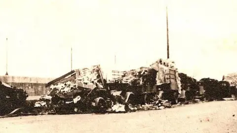 An old sepia toned picture of destroyed lorries on a quayside