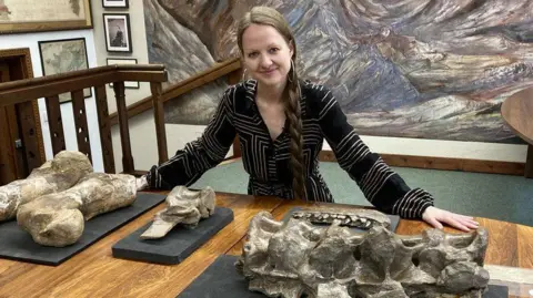 Dr Emma Nicholls and Megalosaurus bones. She is sitting at a desk , with the bones on the table top.