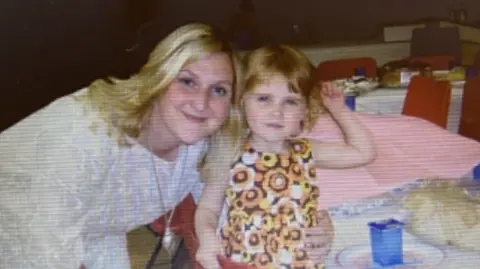 An old picture of me and my mother at my third birthday party. We are both looking at the camera smiling. My mother is on the left and is holding onto me while I am sat at a table. She is wearing a white long sleeved dress. I am on the right. I am wearing a orange, brown and white floral dress. I have short light brown hair.