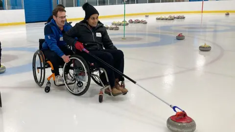 A training session shows a woman in a wheelchair holding a stick touching a curling stone. A man in a blue fleece is behind her in his wheelchair holding her wheels