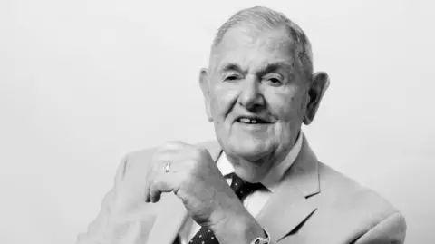 A close up image of Tony Chinn in a black and white portrait photo. He is wearing a suit and tie and smiles, looking beyond the camera.