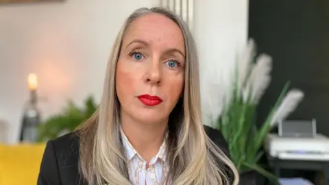 A woman with light coloured long hair looks at a camera. She is smartly dressed. She is in a plain room, sitting next to a tall, green plant. 