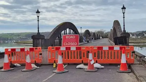 A bridge over the River Dee with a road closed sign and orange barriers and cones across it - houses are visible in the distance