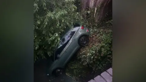 A large grey car that has come off the road and landed underneath an overgrown tunnel. The front of the car is water and the back wheels are stuck on an overgrown verge. Branches from a large tree are covering the roof of the car. A brick wall can be seen above, where the car has fallen from. 