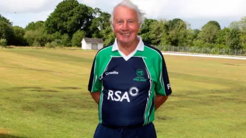 Belfast Telegraph Ossie Colhoun is wearing a dark blue and green cricket Ireland top. He is standing in a green field smiling at the camera.