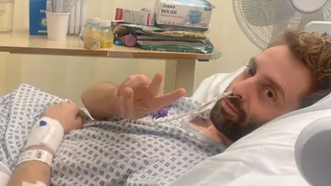 Daniel Harding Daniel Harding is lying in a hospital bed wearing a medical gown. He is holding up his hand in a V sign and smiling slightly. There is a fan behind him and table with a box of facemasks and juice on. 