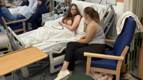 Sophie being comforted by her sister Emily in the cell collection centre as she lies on a bed in the unit 