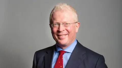 A bespectacled man in a navy suit, blue shirt and red tie.