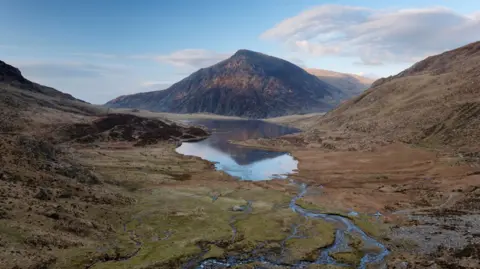 Getty Images A view over Llyn Idwal lake with Pen yr Ole Wen mountain in the distance, at Snowdonia National Park. The green valley is large and wide, with the mountain in the horizon and blue sky with clouds behind. 