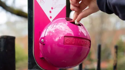 A pink, ball shaped bin on a black railing, with a circular hole on the top. Someone can be seen emptying chewing gum into it 